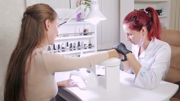 La mujer manicurista en baño cosmético está aplicando gel en las uñas de los clientes. La chica se seca las uñas en una lámpara ultravioleta . — Vídeos de Stock