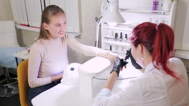 美容室の女性マニキュリストは、クライアントの爪にゲルを適用します。女の子は紫外線ランプで爪を乾かします — ストック動画