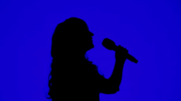 İnce bir kadın şarkıcının silueti mavi arka planda bir mikrofonla sahnede.. — Stok video
