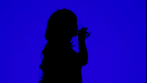 푸른 배경에 있는 유리잔에서 포도주를 마시고 있는 곱슬머리의 여자 실루엣 — 비디오
