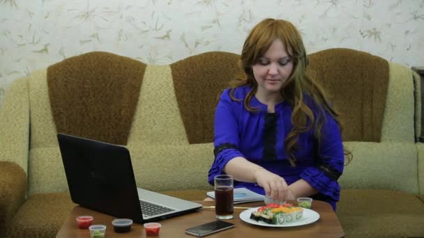 Una joven en una mesa con un portátil come rollos y trabaja — Vídeos de Stock