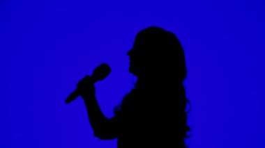 İnce bir kadın şarkıcının silueti mavi arka planda bir mikrofonla sahnede.