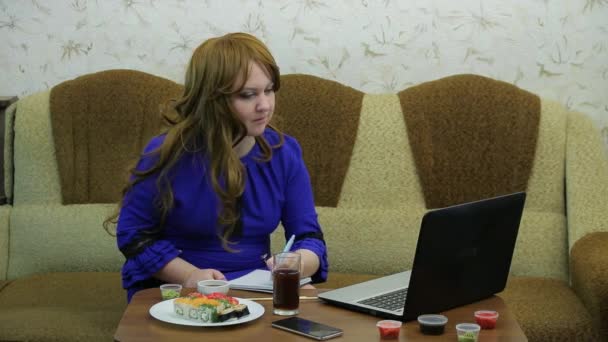 ラップトップを持つテーブルの若い女性がロールと作品を食べる — ストック動画