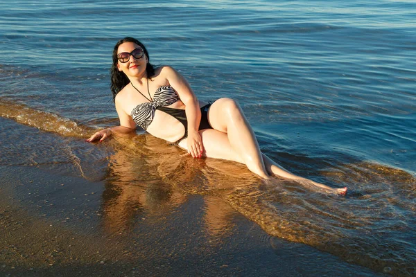 Uma mulher morena em um maiô e óculos está tomando banho de sol na costa . — Fotografia de Stock