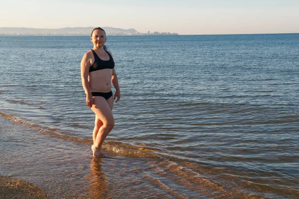 En kvinna i svart baddräkt går längs stranden en solig dag.. — Stockfoto