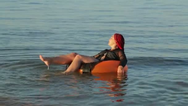 A woman with red hair in a bathing suit on the sea waves in a swimming circle sunbathes — Stock Video