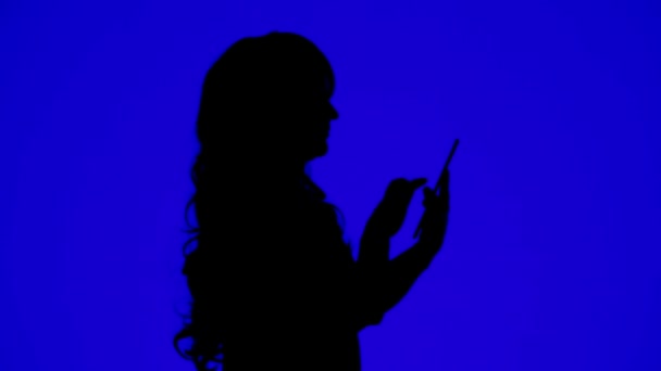 Силуэт тонкой женщины с вьющимися волосами, общающейся в мессенджере на планшете на синем фоне — стоковое видео