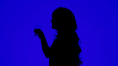 Kıvırcık saçlı, mavi arka planda bardaktan şarap içen bir kadının silüeti.