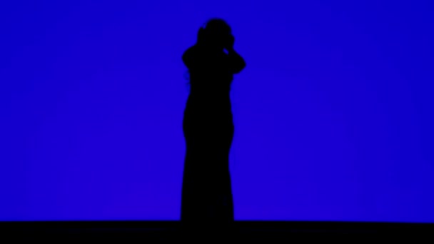 Silhouet van een slanke vrouw in een koptelefoon die danst op de muziek op een blauwe achtergrond — Stockvideo