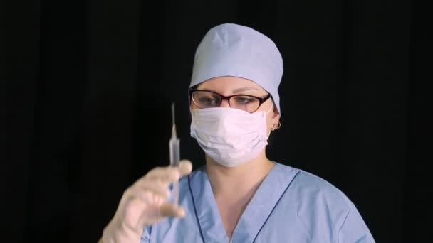 Vrouwelijke arts in een medisch masker met een spuit in de hand op een zwarte achtergrond. — Stockvideo