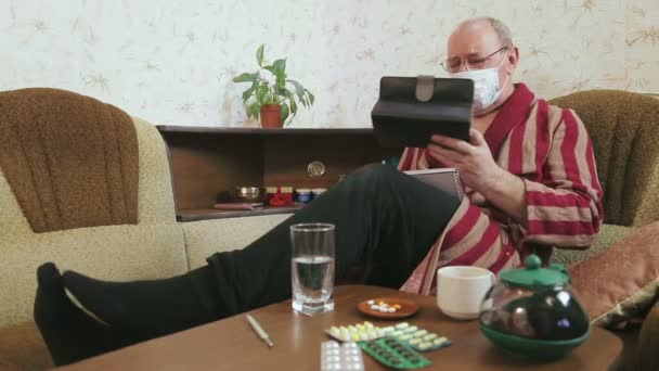 Мужчина в халате на диване дома с симптомами гриппа работает с таблеткой — стоковое видео