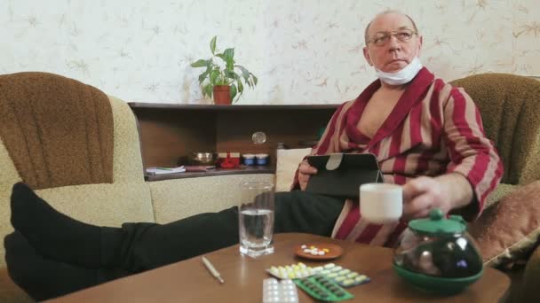Un hombre en un albornoz en cuarentena auto-aislamiento se comunica a través de un enlace de vídeo a través de una tableta en el sofá y bebe té — Vídeos de Stock
