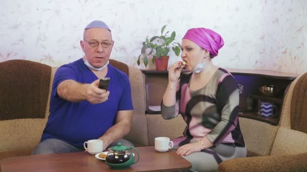 Jewish married couple in protective masks in quarantine for the prevention of coronavirus at home on the couch drinking tea and watching TV — Stock Video