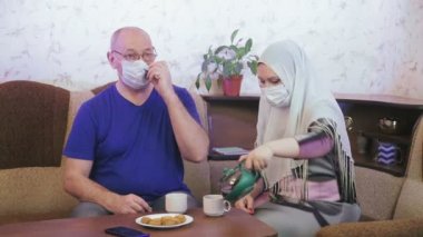 Korunaklı maskeli Müslüman çift evde kanepede çay içip televizyon izleyerek koronavirüsü önlemek için karantinada.