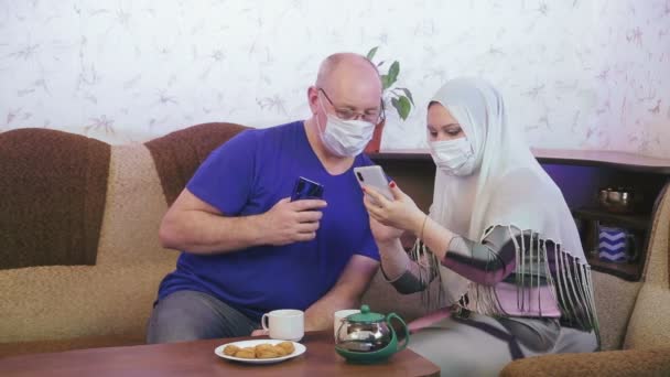 Moslim koppel in beschermende maskers in quarantaine voor de preventie van coronavirus thuis op de bank het nemen van foto 's op een smartphone en het kijken naar foto' s. — Stockvideo