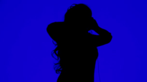 Silhouette of a woman wearing headphones and dancing to the music on a blue background — Stockvideo