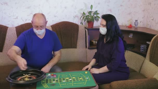 Супружеская пара в медицинских масках играет в рулетку дома в карантине для предотвращения коронавируса — стоковое видео