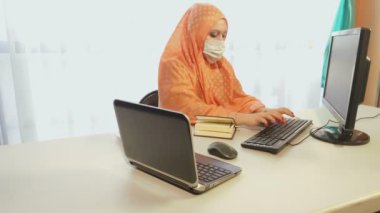 Karantina döneminde tıbbi maske takan Müslüman kadın büroda bilgilerle çalışıyor.