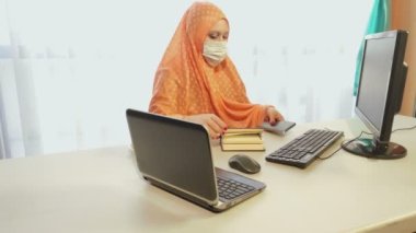 Karantina döneminde tıbbi maske takan Müslüman kadın büroda bilgilerle çalışıyor.