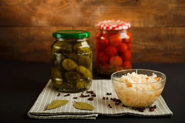 Gläser mit eingelegten Tomaten und Gurken und Sauerkraut auf einem Holztisch vor schwarzem Hintergrund. — Stockfoto