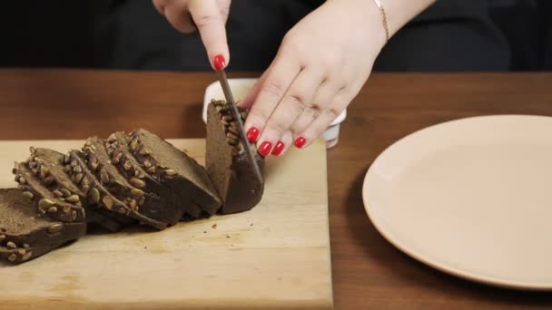 Uma mão feminina corta com uma faca em uma tábua de madeira pão de centeio escuro com farelo de cereais — Vídeo de Stock