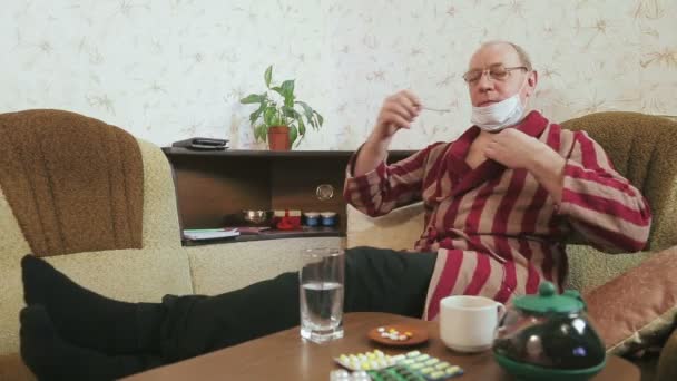 Mężczyzna w szlafroku na kanapie w domu z objawami grypy mierzy temperaturę. — Wideo stockowe