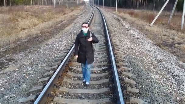 Koruyucu sağlık maskesi takmış yorgun bir kadın tren yolu boyunca elinde bavulla yürüyor. — Stok video