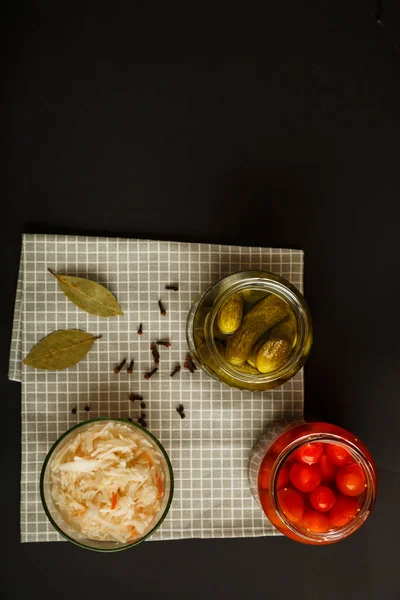 Burkar med inlagda tomater och gurkor och surkål på ett träbord på en svart bakgrund. — Stockfoto