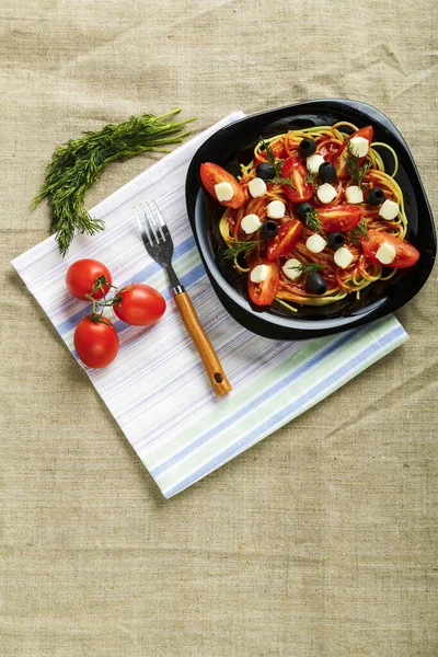 Un plato negro con espaguetis en una salsa decorada con tomates cherry aceitunas y mozzarella en una servilleta sobre un mantel de lino . —  Fotos de Stock