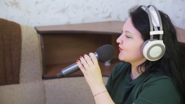 Женщина дома на диване в наушниках поет в караоке микрофон — стоковое видео