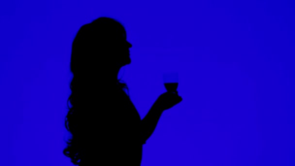 Egy göndör hajú nő sziluettje, aki egy kék hátterű pohárból iszik bort — Stock videók