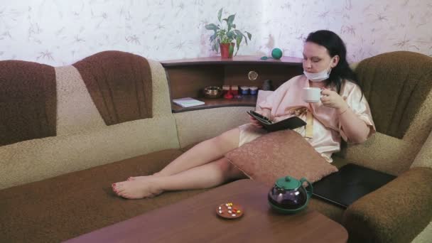 一名身穿家居服、戴着医疗面罩、戴着隔离隔离装置的年轻女子在沙发上喝茶，并在平板电脑上看书. — 图库视频影像