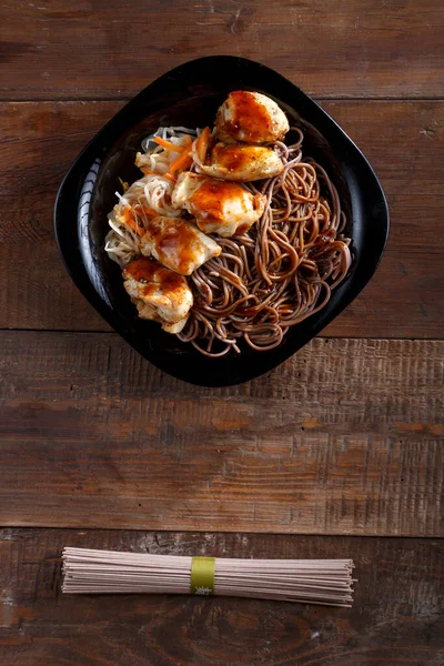 Zwarte plaat met soba noedels met kip en groenten in teriyaki saus op een lichte houten tafel. — Stockfoto