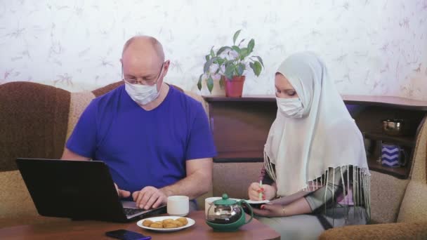 Casal muçulmano em máscaras protetoras em quarentena para a prevenção de coronavírus em casa no sofá envolvido em trabalho remoto — Vídeo de Stock