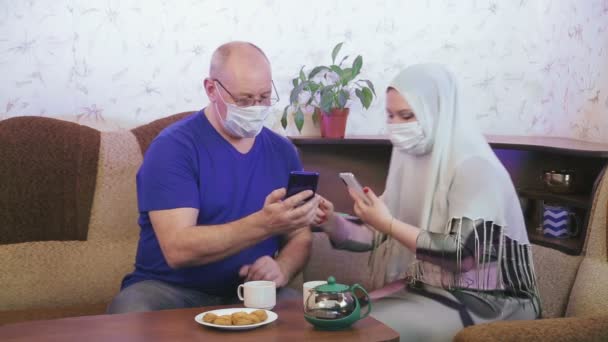スマートフォンで写真を撮ると写真を見てソファの上に自宅でコロナウイルスの予防のために隔離された保護マスクのイスラム教徒のカップル — ストック動画