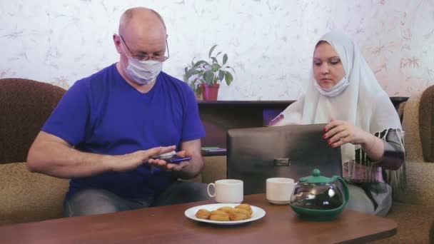 Muslimisches Paar in Schutzmasken in Quarantäne zur Vorbeugung gegen Coronavirus zu Hause auf der Couch desinfizieren mit Feuchttüchern Hände und Smartphones und Laptop — Stockvideo