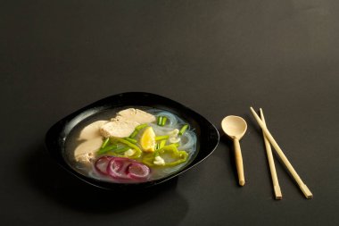 Siyah arka planda tavuklu bir tabak Vietnamlı Pho çorbası, kaşık ve çubuklarla..