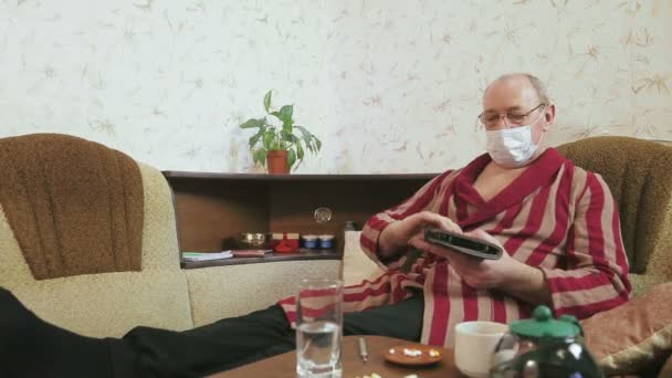 Человек в домашнем пальто и медицинская маска в карантинной самоизоляции работает на планшете на диване . — стоковое видео