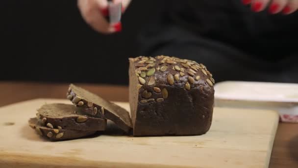 A female hand slices with a knife on a wooden board dark rye bread with bran. — Stock Video