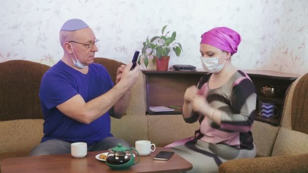 Еврейская супружеская пара в защитных масках в карантине для предотвращения коронавируса дома на диване фотографируются в масках — стоковое видео