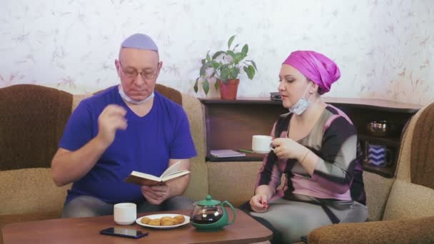 Ζευγάρι Εβραίων με προστατευτικές μάσκες σε καραντίνα για την πρόληψη του κορωνοϊού στο σπίτι στον καναπέ σύζυγο που διαβάζει ένα βιβλίο και πίνει τσάι. — Αρχείο Βίντεο
