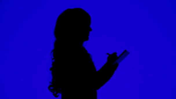 Silhouette di una donna snella con i capelli ricci che scrive con una matita in un quaderno su uno sfondo blu . — Video Stock