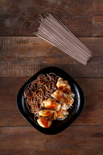 Zwarte plaat met soba noedels met kip en groenten in teriyaki saus op een lichte houten tafel. — Stockfoto