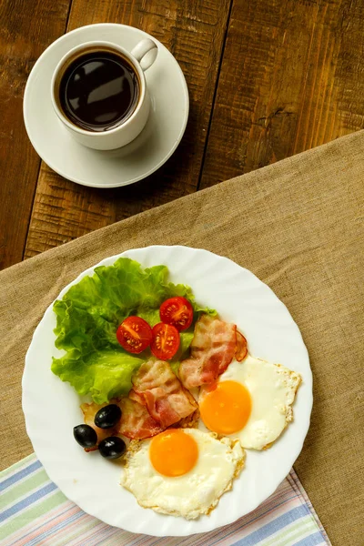 En una mesa de madera hay un plato con huevos fritos y una taza de café . — Foto de Stock