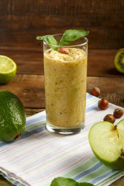 Bir bardak yeşil smoothie, ahşap bir masada, bir peçetenin üzerinde, avokado elmalı limonlu fındıkların yanında..