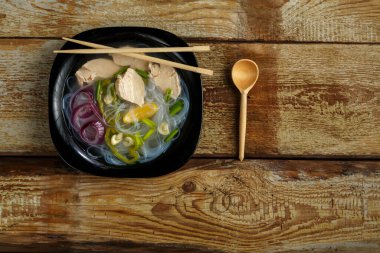 Tahta bir masa ve aletlerin üzerinde tavuk olan siyah bir tabak Vietnam Pho çorbası..