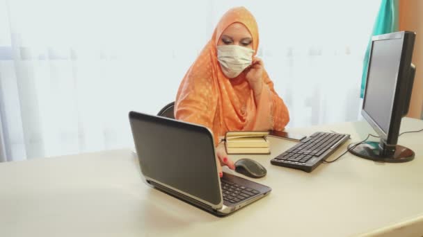 Moslim vrouw in hijab in een medisch masker tijdens de quarantaine periode werkt in het kantoor met gadgets. — Stockvideo