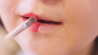 Genç bir kadın yarım dudağına aynanın önünde fırçayla kırmızı ruj sürüyor..