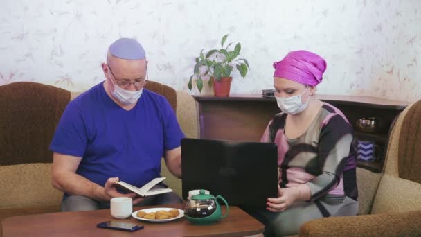 Couple marié juif en masques protecteurs mis en quarantaine pour la prévention du coronavirus à la maison sur le canapé mari lit un livre femme travaille sur l'ordinateur . — Video