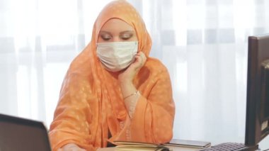 Ofiste koruyucu maskeli Müslüman bir kadın bilgisayar başında ve karantina döneminde bilgi ile çalışıyor..